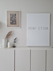 A minimalist interior scene with a neutral color palette. On the wall, two pieces of framed artwork are displayed: one featuring abstract vertical shapes, and the other with the incomplete word 'PERFECTION' missing the letter 'E'. Below the frames, there is a white cabinet, decorated with two white vases - one holding dried pampas grass, and a small black abstract sculpture.