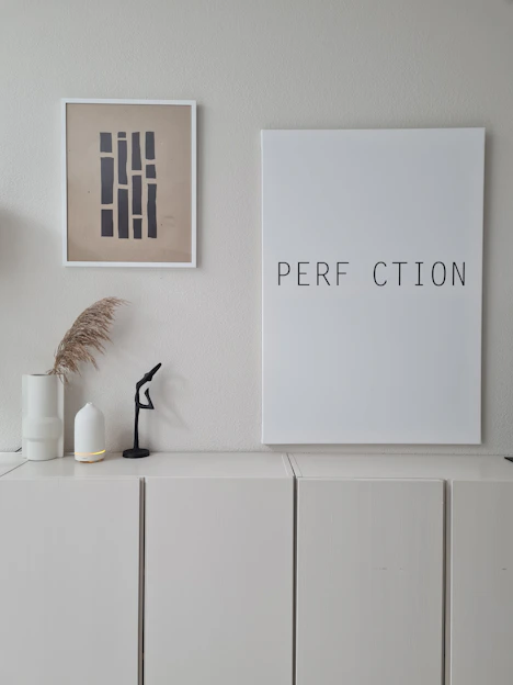 A minimalist interior scene with a neutral color palette. On the wall, two pieces of framed artwork are displayed: one featuring abstract vertical shapes, and the other with the incomplete word 'PERFECTION' missing the letter 'E'. Below the frames, there is a white cabinet, decorated with two white vases - one holding dried pampas grass, and a small black abstract sculpture.