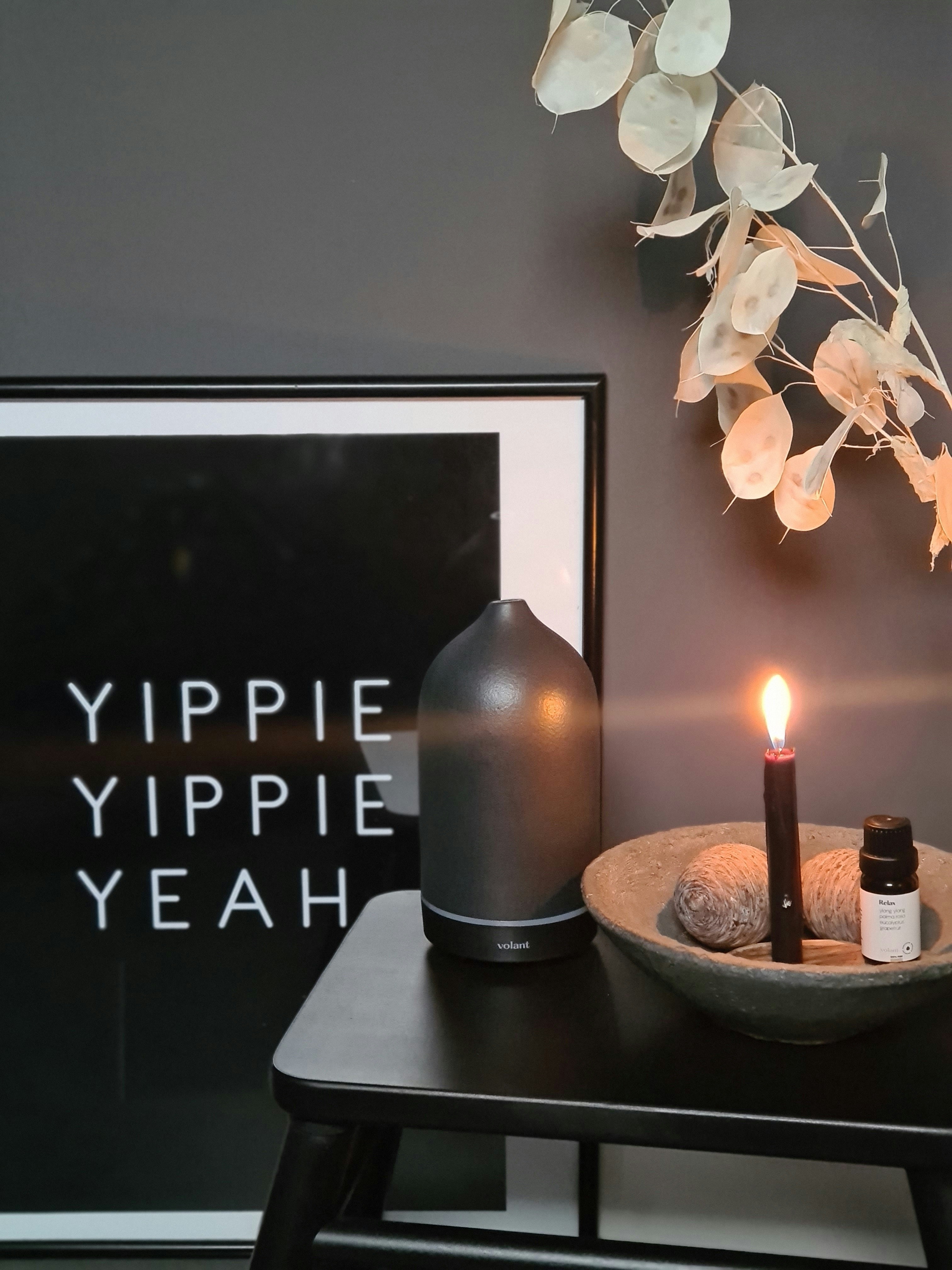 Luxury scented candle and diffuser in dark minimalist interior, warm amber glow, atmospheric luxury lifestyle setting with botanical elements