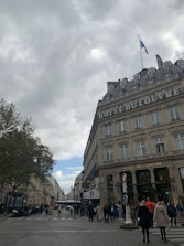 A bustling street scene with people walking in a city. The image features a historic building with the words 'HOTEL DU LOUVRE' prominently displayed. A French flag flies atop the building, and trees line the street. The sky is overcast with patches of blue visible.