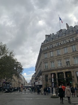 A bustling street scene with people walking in a city. The image features a historic building with the words 'HOTEL DU LOUVRE' prominently displayed. A French flag flies atop the building, and trees line the street. The sky is overcast with patches of blue visible.