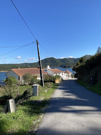 A rural street lined with a few white houses with red roofs. A telephone pole and power lines run alongside the road, leading towards a body of water and forested hills in the distance. The sky is clear blue, suggesting a sunny day.