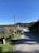 A rural street lined with a few white houses with red roofs. A telephone pole and power lines run alongside the road, leading towards a body of water and forested hills in the distance. The sky is clear blue, suggesting a sunny day.