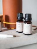 Several small brown bottles with black caps are placed on a light surface near a large orange cylindrical diffuser. The labels on the bottles describe different essential oil blends, such as 'Energy', indicating scents like lime and grapefruit. A white pen is visible in the foreground.