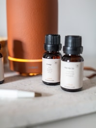 Several small brown bottles with black caps are placed on a light surface near a large orange cylindrical diffuser. The labels on the bottles describe different essential oil blends, such as 'Energy', indicating scents like lime and grapefruit. A white pen is visible in the foreground.