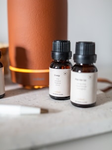 Several small brown bottles with black caps are placed on a light surface near a large orange cylindrical diffuser. The labels on the bottles describe different essential oil blends, such as 'Energy', indicating scents like lime and grapefruit. A white pen is visible in the foreground.