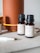 Several small brown bottles with black caps are placed on a light surface near a large orange cylindrical diffuser. The labels on the bottles describe different essential oil blends, such as 'Energy', indicating scents like lime and grapefruit. A white pen is visible in the foreground.