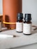 Several small brown bottles with black caps are placed on a light surface near a large orange cylindrical diffuser. The labels on the bottles describe different essential oil blends, such as 'Energy', indicating scents like lime and grapefruit. A white pen is visible in the foreground.