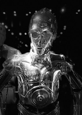 A sleek, metallic humanoid robot with a shiny surface reflecting ambient light. It has large, round eyes and detailed mechanical features, set against a dark background with scattered bright spots.