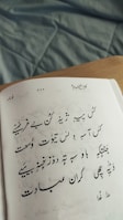 A page from an Urdu poetry book with handwritten text in elegant calligraphy. The paper shows signs of shadow and texture, resting on a wooden surface with a crumpled fabric background in light blue.