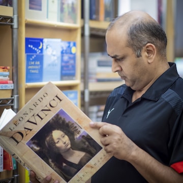 A man is engrossed in reading a large book about Leonardo Da Vinci in a bookstore. Shelves filled with various books are visible in the background, suggesting a rich literary environment. The man is wearing a black shirt and appears to be in deep concentration.