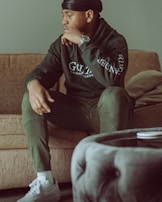 A person sits thoughtfully on a textured sofa with an arm resting on a cushion. Dressed in casual attire including a dark hoodie with lettering and matching pants, they wear a watch on the wrist and white sneakers. The setting is a softly lit room with neutral colors, and a round upholstered piece of furniture appears in the foreground.