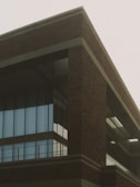 A building with a modern architectural design featuring large glass windows and brickwork. The structure is prominently angular with multiple floors visible. The lighting suggests an overcast sky, contributing to a muted atmosphere.