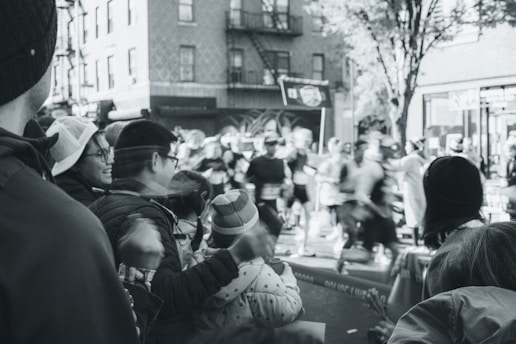 A black and white photograph captures a lively street scene during a marathon or race. In the foreground, people are gathered, cheering and watching the race. Some individuals appear to be taking photos or videos of the event. In the background, blurred motion of the runners can be seen as they pass by buildings and trees, emphasizing the pace and energy of the race. The image has an overall festive and energetic atmosphere.