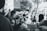 A black and white photograph captures a lively street scene during a marathon or race. In the foreground, people are gathered, cheering and watching the race. Some individuals appear to be taking photos or videos of the event. In the background, blurred motion of the runners can be seen as they pass by buildings and trees, emphasizing the pace and energy of the race. The image has an overall festive and energetic atmosphere.