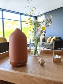 A modern and minimalistic interior scene featuring a wooden tray on a table. On the tray, there is a terracotta-colored diffuser, a transparent glass vase with assorted wildflowers such as daisies and yellow blooms, and a small gold decorative object with a curved handle. In the background, there is a living room with a dark-colored couch and mustard-yellow cushions against a blue wall. Large windows let in natural light and provide a view of greenery outside.