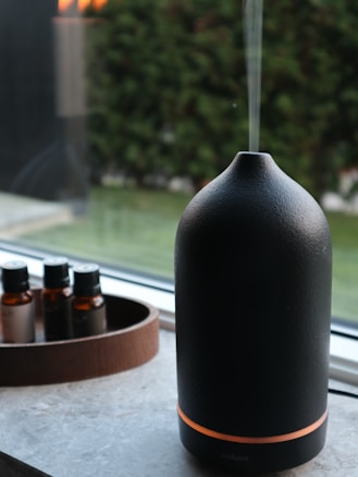 A sleek, dark-colored essential oil diffuser emits a thin stream of vapor, placed on a window sill with a view of a blurred garden. Next to it is a wooden tray holding small, dark glass bottles of essential oils.