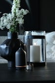 A dark-toned setting featuring a black geometric vase holding delicate white flowers. Nearby, a black essential oil diffuser emits a soft mist. A large white candle sits inside a glass holder, with a few small bottles of essential oils placed in the foreground.
