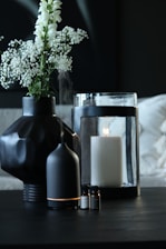 A dark-toned setting featuring a black geometric vase holding delicate white flowers. Nearby, a black essential oil diffuser emits a soft mist. A large white candle sits inside a glass holder, with a few small bottles of essential oils placed in the foreground.