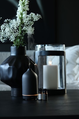 A dark-toned setting featuring a black geometric vase holding delicate white flowers. Nearby, a black essential oil diffuser emits a soft mist. A large white candle sits inside a glass holder, with a few small bottles of essential oils placed in the foreground.