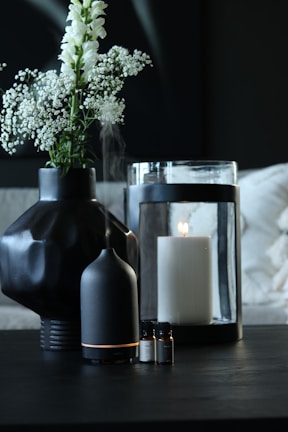 A dark-toned setting featuring a black geometric vase holding delicate white flowers. Nearby, a black essential oil diffuser emits a soft mist. A large white candle sits inside a glass holder, with a few small bottles of essential oils placed in the foreground.