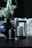 A dark-toned setting featuring a black geometric vase holding delicate white flowers. Nearby, a black essential oil diffuser emits a soft mist. A large white candle sits inside a glass holder, with a few small bottles of essential oils placed in the foreground.