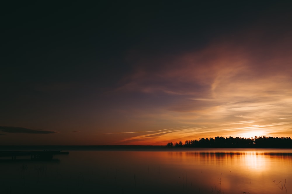 the sun is setting over a lake with a bench in the foreground