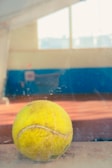 A close-up of a used tennis ball resting on a reflective surface with an indoor tennis court in the blurred background. The ball shows signs of wear and is placed in the foreground, while natural light floods through large windows.