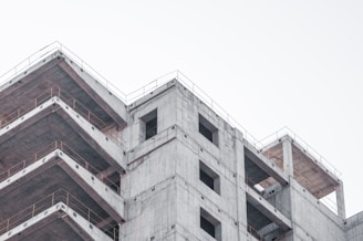 A partially constructed concrete building with multiple floors, exposed rebar, and scaffolding around the edges. The sky is overcast, creating a muted background.