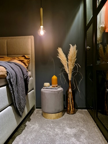 A cozy bedroom corner with a modern aesthetic features a light grey upholstered bed. A pendant light with a vintage bulb softly illuminates the space, casting a warm glow. Beside the bed is a chic grey stool with a golden base, topped with a stack of books and an amber-colored candle. A tall vase filled with dried pampas grass adds a touch of natural texture and elegance. The carpet is a subtle grey, complementing the dark walls, while a mirrored wardrobe adds depth to the room.