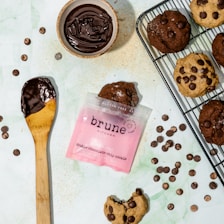 A variety of chocolate chip cookies are arranged on a cooling rack and a light-colored surface. A bowl of melted chocolate and a wooden spoon with chocolate are nearby. There's a pink package labeled 'brune kitchen, double chocolate chip cookie'. Chocolate chips are scattered across the surface.