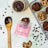A variety of chocolate chip cookies are arranged on a cooling rack and a light-colored surface. A bowl of melted chocolate and a wooden spoon with chocolate are nearby. There's a pink package labeled 'brune kitchen, double chocolate chip cookie'. Chocolate chips are scattered across the surface.