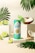 A turquoise and green energy drink can labeled 'Positive Energy' stands on a white platform. It is surrounded by fresh coconut halves and lime slices, with tropical palm leaves adding to the vibrant, tropical theme. A small green and white paper umbrella is placed inside a coconut half, enhancing the tropical atmosphere.