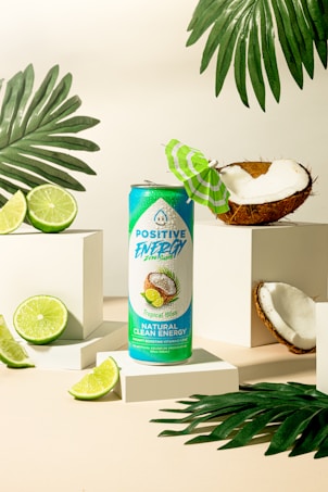 A turquoise and green energy drink can labeled 'Positive Energy' stands on a white platform. It is surrounded by fresh coconut halves and lime slices, with tropical palm leaves adding to the vibrant, tropical theme. A small green and white paper umbrella is placed inside a coconut half, enhancing the tropical atmosphere.