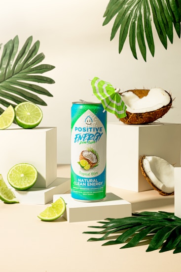 A turquoise and green energy drink can labeled 'Positive Energy' stands on a white platform. It is surrounded by fresh coconut halves and lime slices, with tropical palm leaves adding to the vibrant, tropical theme. A small green and white paper umbrella is placed inside a coconut half, enhancing the tropical atmosphere.