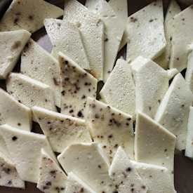 Several pieces of a white, porous snack have been cut into irregular shapes and arranged on a surface. Some pieces are plain, while others are speckled with what looks like small black seasoning or peppercorns.