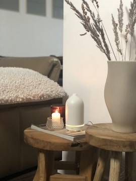 A cozy setting with a lit candle in a glass holder placed on a wooden stool. Next to it is a white ceramic diffuser and a stack of magazines. A large beige vase containing dried grasses adds a natural element. In the background, there's a comfortable, cushioned seat with a textured cover.