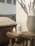 A cozy setting with a lit candle in a glass holder placed on a wooden stool. Next to it is a white ceramic diffuser and a stack of magazines. A large beige vase containing dried grasses adds a natural element. In the background, there's a comfortable, cushioned seat with a textured cover.