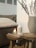 A cozy setting with a lit candle in a glass holder placed on a wooden stool. Next to it is a white ceramic diffuser and a stack of magazines. A large beige vase containing dried grasses adds a natural element. In the background, there's a comfortable, cushioned seat with a textured cover.