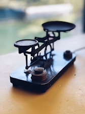A vintage mechanical balance scale with two platforms is placed on a light-colored surface. The scale has a rustic, metallic appearance, featuring visible weights and intricate components. The background is softly blurred, suggesting an indoor setting with natural light coming from a window.