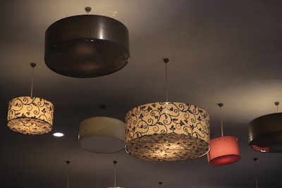 A ceiling adorned with multiple hanging lampshades, each with unique designs and colors. The lampshades have intricate patterns, including floral and swirl motifs, and vary in color from dark brown to beige and pink.