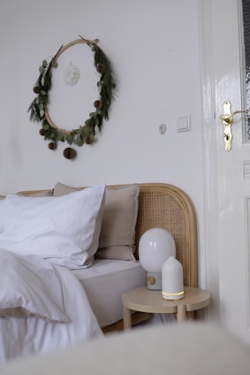 A cozy bedroom corner features a wicker bed frame with white and beige bedding. Above the bed, a delicate decorative wreath made of leaves and small fluffy elements hangs on the wall. Two small wooden tables beside the bed hold a modern white table lamp and a small diffuser. The room has a soft and minimalist aesthetic.