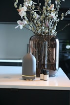 A modern interior setting with a vase containing branches and flowers placed on a countertop. Beside the vase, there is a sleek, cylindrical diffuser with essential oil bottles next to it.