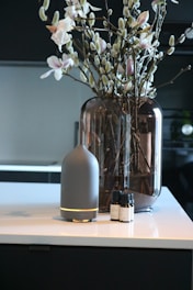 A modern interior setting with a vase containing branches and flowers placed on a countertop. Beside the vase, there is a sleek, cylindrical diffuser with essential oil bottles next to it.