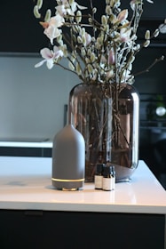 A modern interior setting with a vase containing branches and flowers placed on a countertop. Beside the vase, there is a sleek, cylindrical diffuser with essential oil bottles next to it.