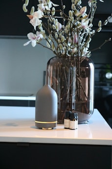 A modern interior setting with a vase containing branches and flowers placed on a countertop. Beside the vase, there is a sleek, cylindrical diffuser with essential oil bottles next to it.