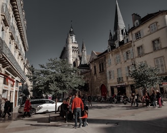 A bustling city street lined with historic buildings and a prominent church tower. People are gathered around benches under trees, with some walking along the street. A white car is parked nearby, and various shops are visible in the background.
