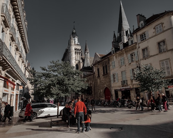 A bustling city street lined with historic buildings and a prominent church tower. People are gathered around benches under trees, with some walking along the street. A white car is parked nearby, and various shops are visible in the background.