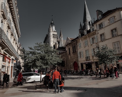 A bustling city street lined with historic buildings and a prominent church tower. People are gathered around benches under trees, with some walking along the street. A white car is parked nearby, and various shops are visible in the background.