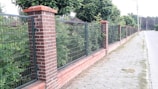 A brick and metal fence runs alongside a sidewalk lined with trees. The fence consists of alternating sections of brick pillars and metal mesh panels. Behind the fence, lush green foliage is visible, and the sidewalk is made of light gray pavers. The scene appears to be in a suburban or residential area.